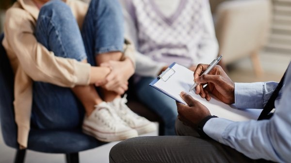 Close-up of a health professional writing on a clipboard while talking to a young person with their knees drawn up to their chest