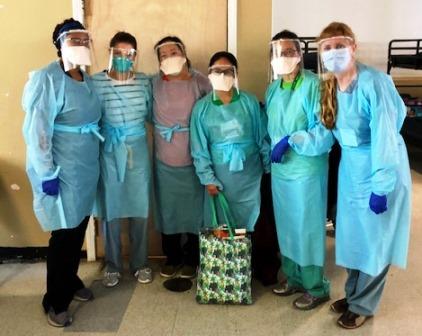 Six faculty members in scrubs and full face masks gather indoors.