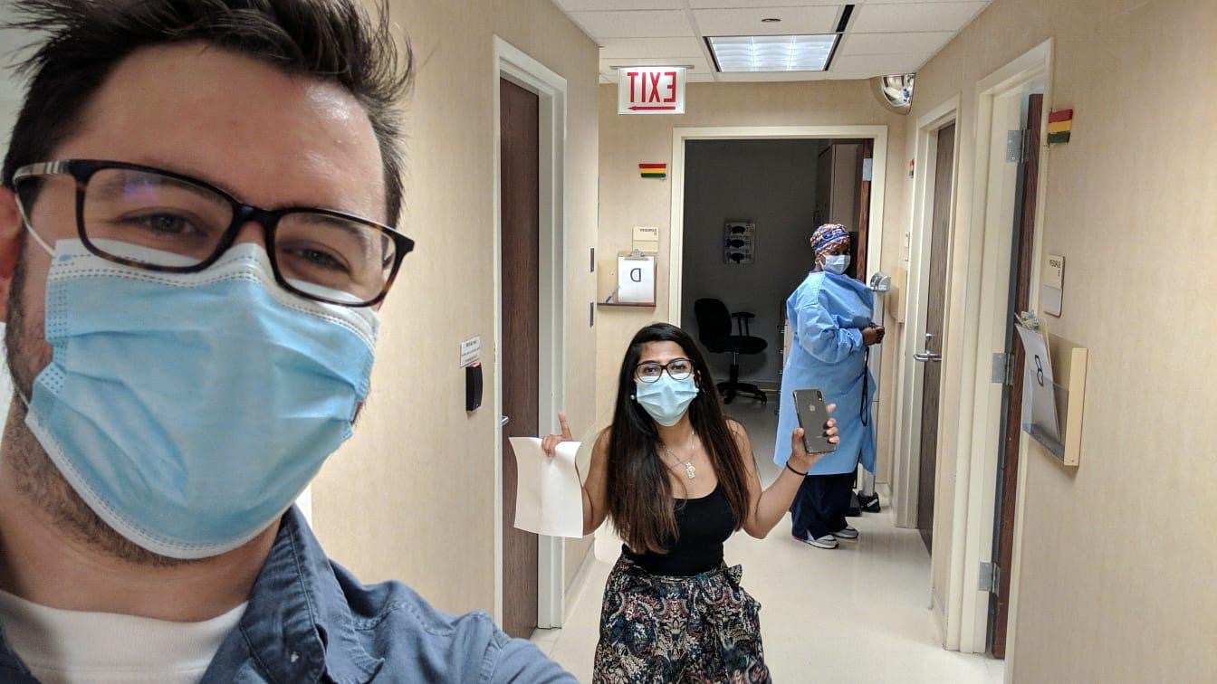 A fellow takes a selfie wearing a mask in the hospital hallway. There is another fellow in a mask in the background posing with a cell phone in one hand and a document in the other. Behind her, a physician in scrubs enters a room attached to the hallway.