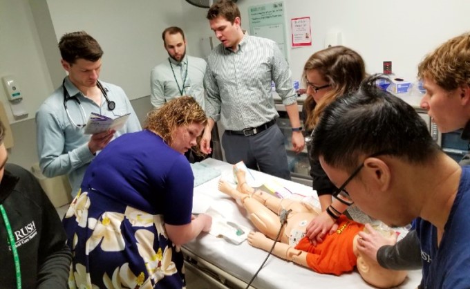 A group of fellows surround a child sized mannequin on an exam table in a hospital room. Two of the fellows are demonstrating CPR on the mannequin.
