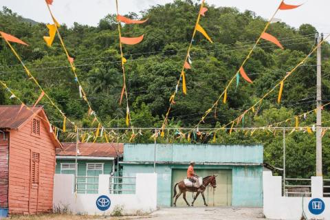 A person rides a horse on a ranch. There are strings of decorative flags stretching from the roof of nearby ranch buildings.