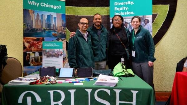 Four people wearing RUSH gear stand at a RUSH booth highlighting the school's diversity, equity and inclusion efforts.