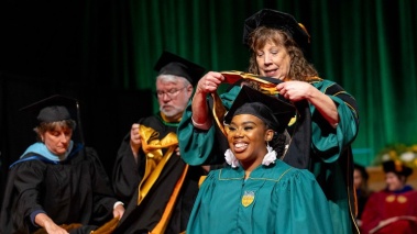 A faculty member wearing academic regalia places a sash over the head of a smiling graduate
