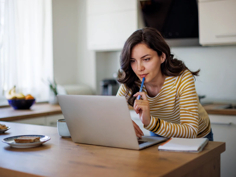 A woman stands in a kitchen, leaning forward to use a laptop resting on a wooden countertop and holding a pen to her lips