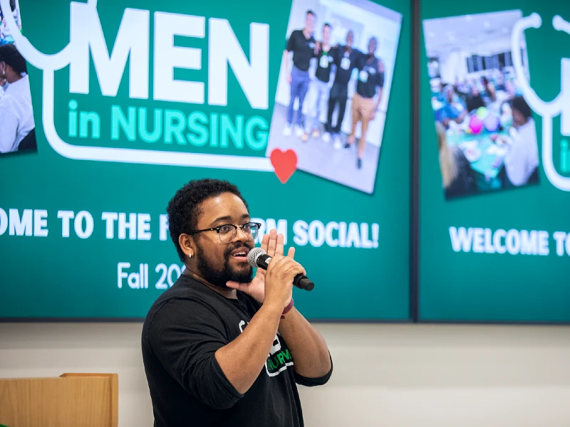 A speaker holds a microphone in front of a presentation reading Men in Nursing