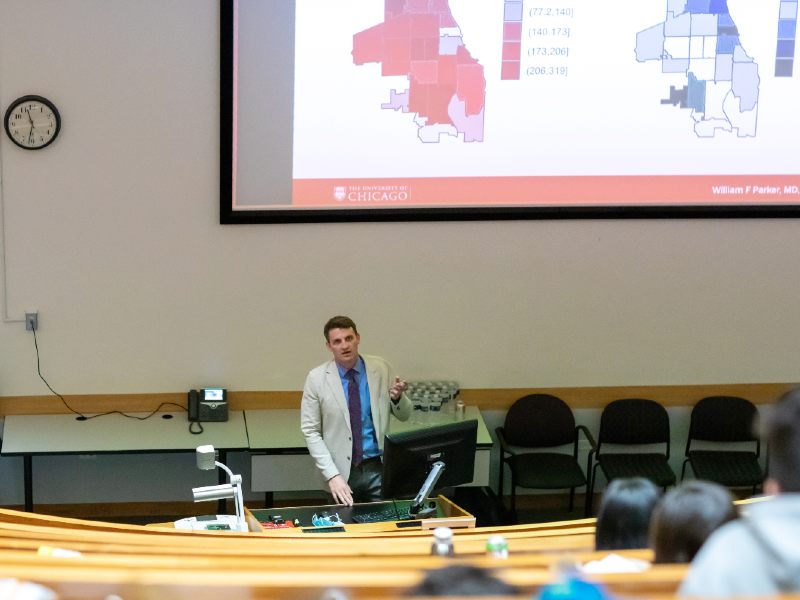 A speaker addresses an audience in a lecture room, with maps of Chicago on the overhead screen behind him