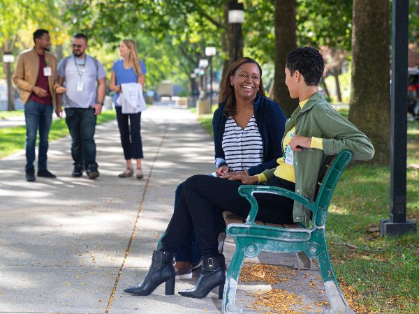 Two students seated on an outdoor bench with others walking in the background