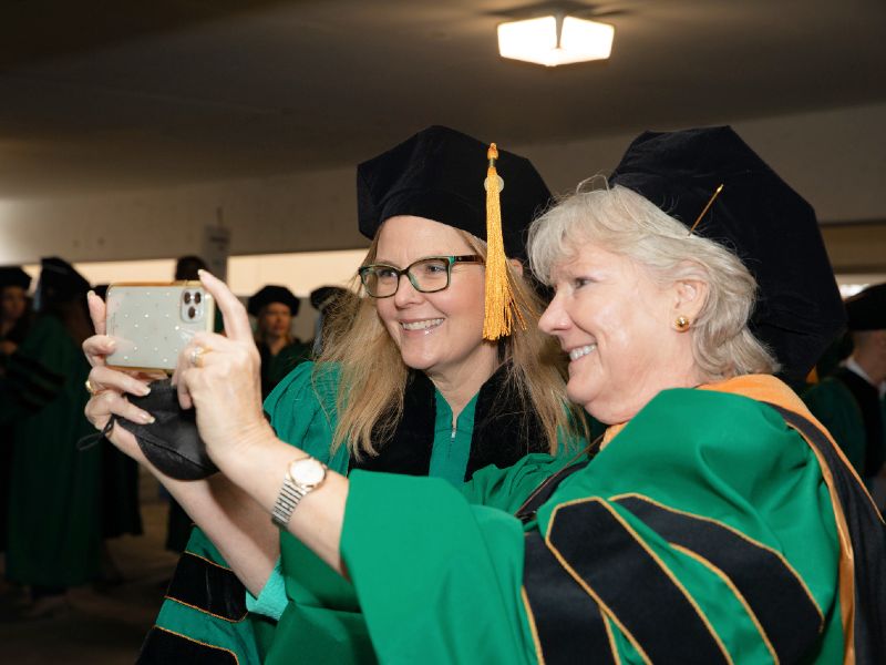 Two faculty members stand together to take a selfie at graduation