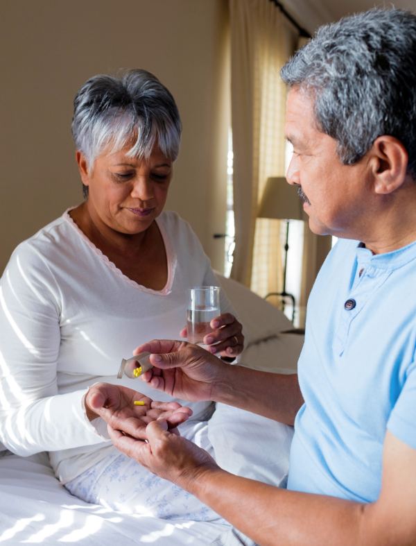 A woman sits up in bed holding a glass of water while a man hands her medication from a pill bottle