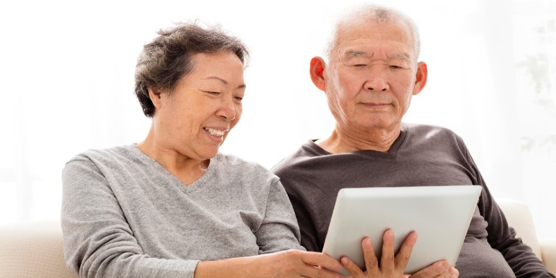 A smiling older man and woman seated on a couch look at a tablet