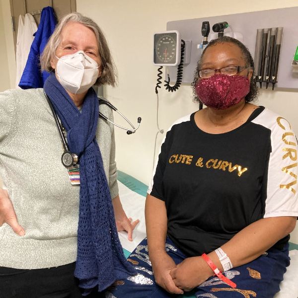 A health care provider stands beside a seated patient, both wearing protective face masks
