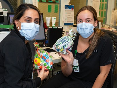 Two seated nurses wearing scrubs and masks hold up colorful care packages