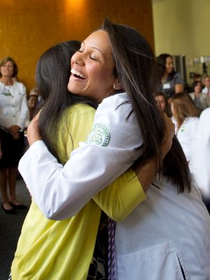 Two women, one wearing a white coat, lean close to hug each other
