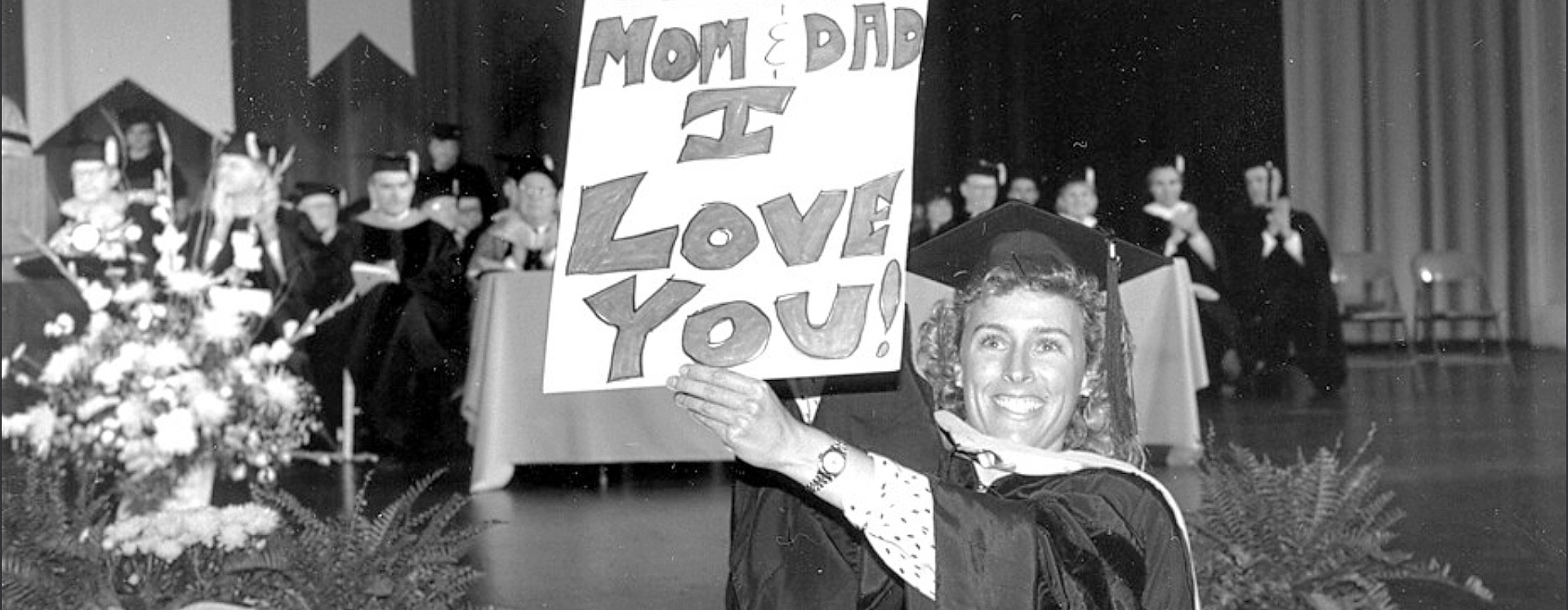 Archival photo of a smiling graduate on stage holding a sign that reads Mom and Dad I Love You