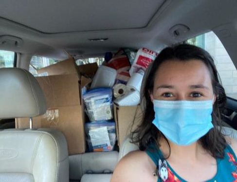 A woman wearing a surgical mask sits in the driver's seat of a car, with boxes of supplies visible in the back seat