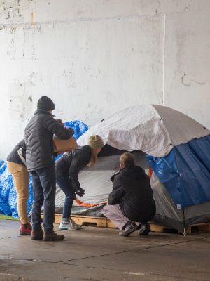 Students carrying supplies kneel at the entrance to a tent beneath a highway overpass