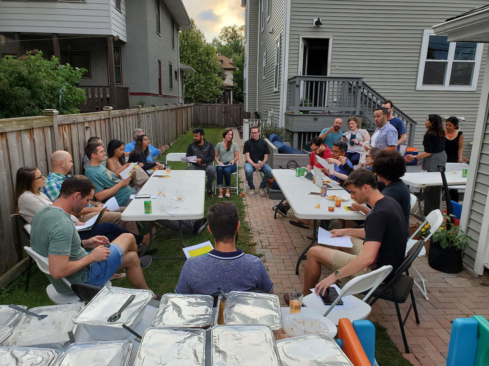 Members sit at two tables laid out in a backyard. There is catered food in the foreground.