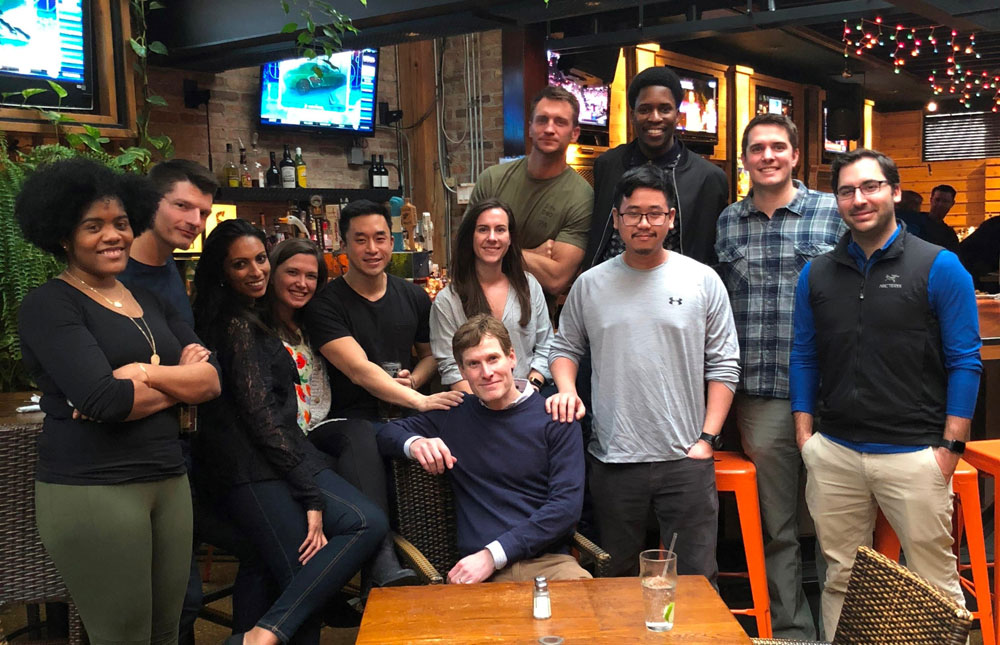 The class of 2020 poses in casual clothing in a restaurant with brick walls and televisions in the background.