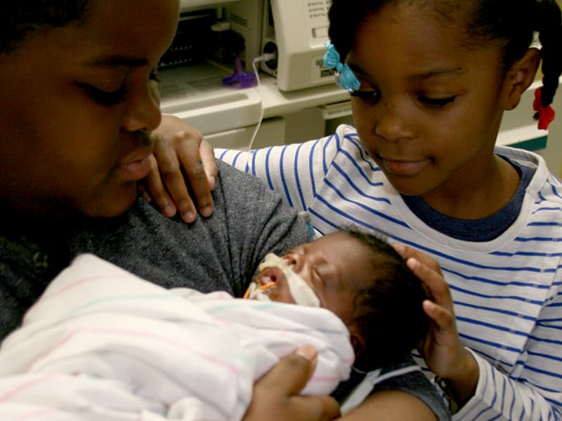 A parent and young child comfort a baby receiving support at the NICU.