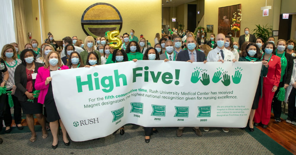 RUSH staff members stand in a gathering space. Several hold a sign that says "High Five" with additional text about RUSH's fifth Magnet Designation.
