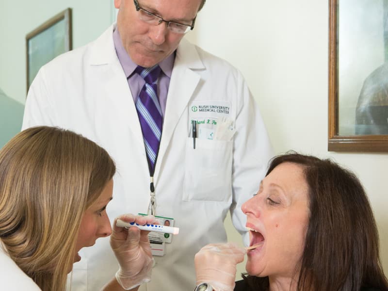 A doctor uses a tongue depressor and flashlight to look at a patient's mouth while another doctor looks on.