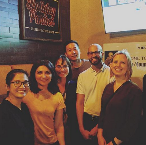 Six third year fellows wear casual clothes and stand inside at a social gathering. The sign on the brick wall behind them reads "Holiday Parties".