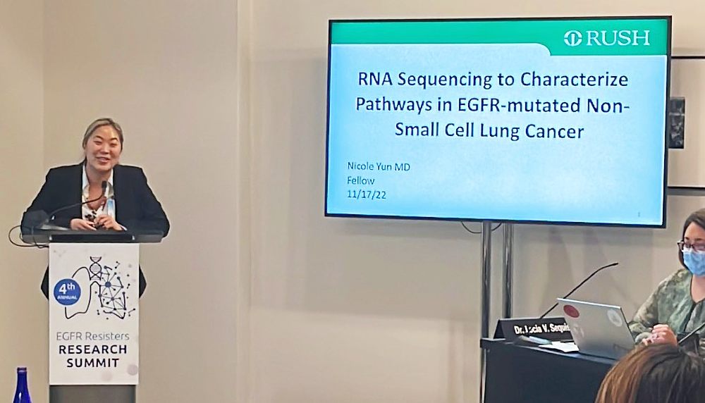 Dr. Yun stands at a podium that says "Research Summit" next to a monitor that reads "RNA Sequencing to Characterize Pathways..."