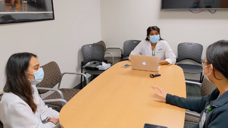 Three people in face masks and lab coats sit at a conference room table and have a discussion. One is taking notes on a laptop.