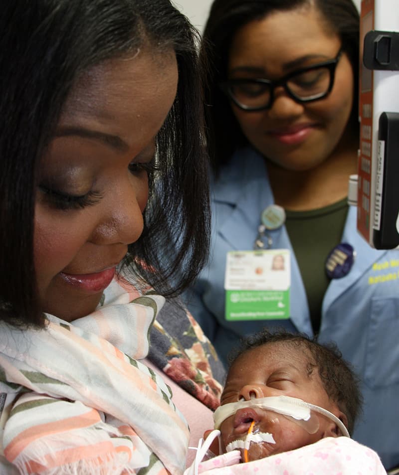 A mother smiles and holds her intubated baby as a physician looks on and smiles in the background.