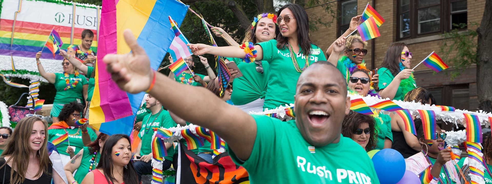 People wearing green RUSH shirts ride a parade float waving rainbow flags. A man in the foreground opens his arm in a welcoming gesture.
