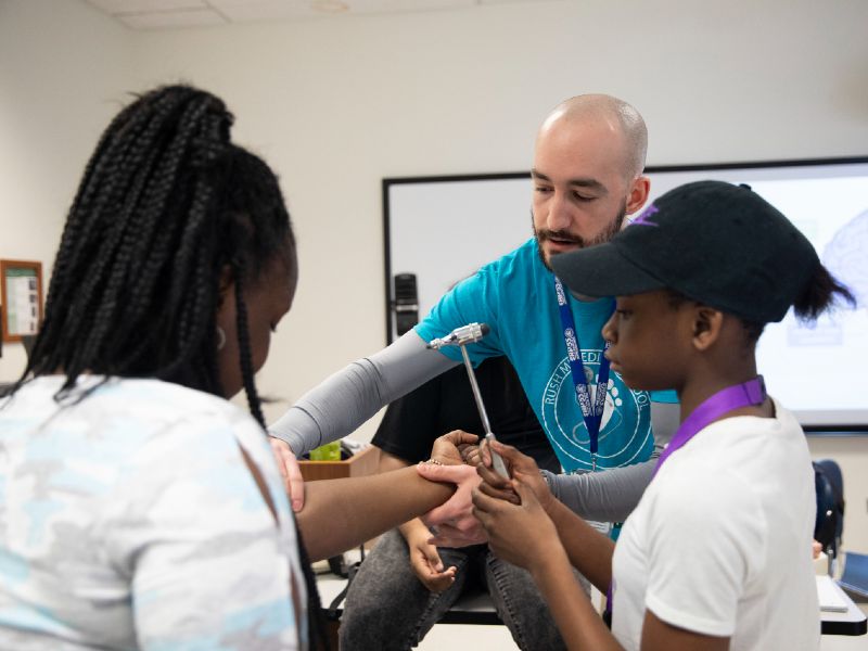 A medical student shows two elementary school students how to test reflexes