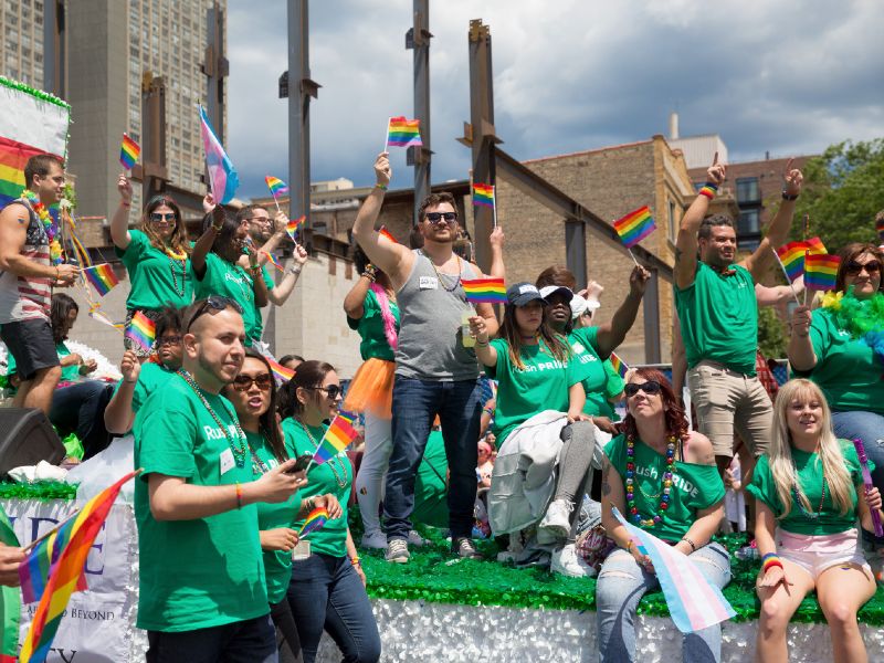 People wearing Rush shirts and waving rainbow flags ride on and walk beside a parade float