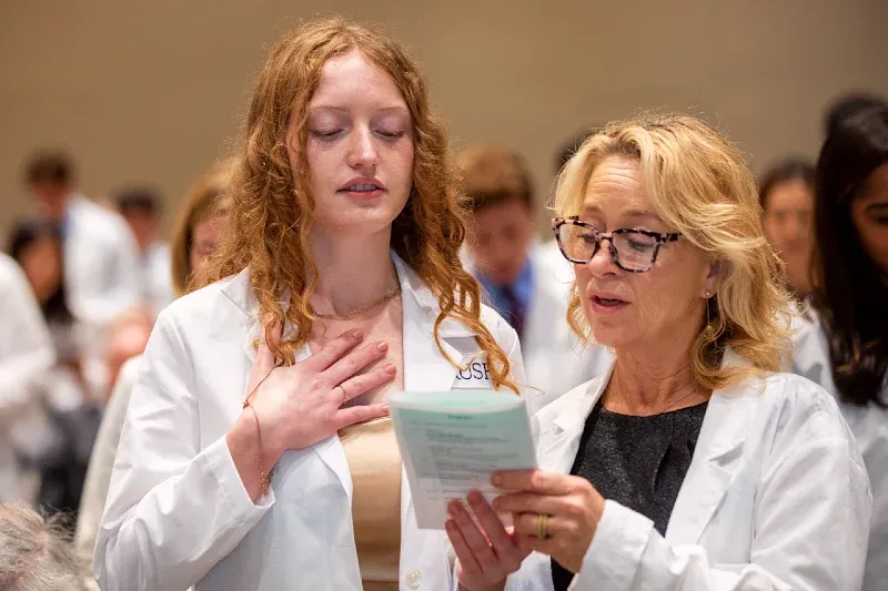 A student wearing a white coat holds her hand over her heart and reads an oath from a piece of paper