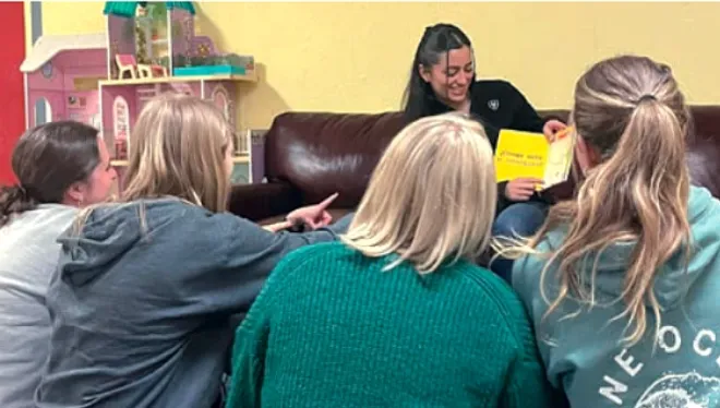 A woman sits on a sofa, reading from a book while showing the pages to a group of girls seated on the floor