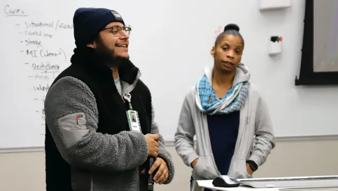 Two people stand in front of a whiteboard speaking to a class