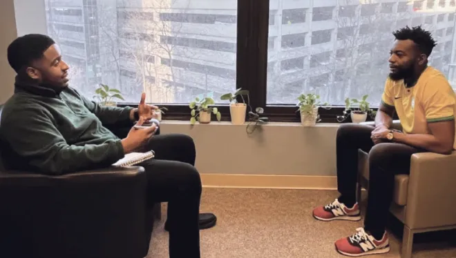 Two men seated in chairs facing one another, with windows behind them and potted plants on the windowsill