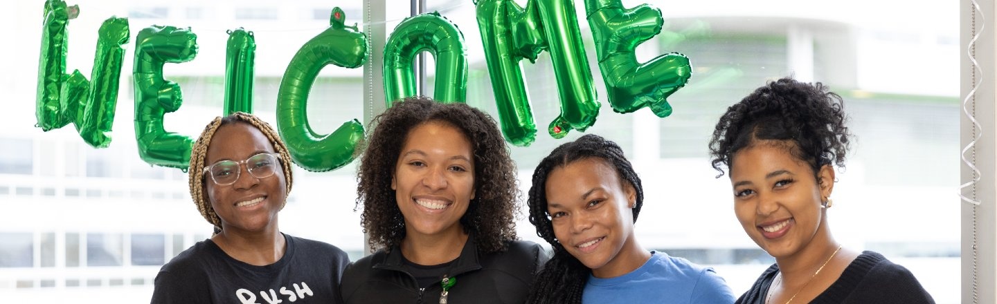 Four students smiling beneath a banner of balloons spelling out Welcome