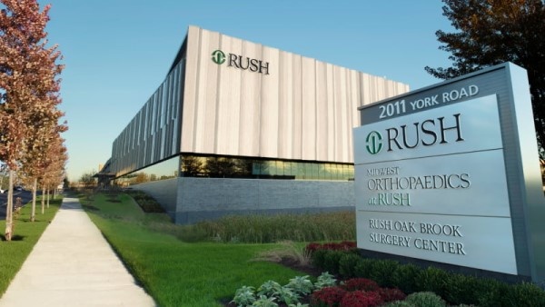 Exterior of RUSH Oak Brook against a clear blue sky. The grass is green and the leaves on the trees are turning reddish brown for fall. A sign in front shows the building address.