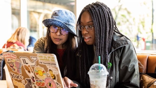 Two young adults sit in a cafe looking at a laptop screen