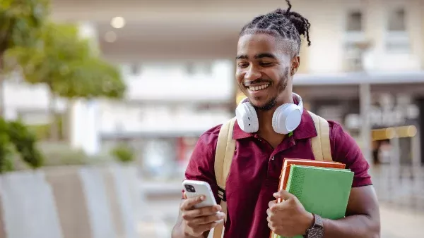 A student wearing headphones, carrying books and looking at his phone screen