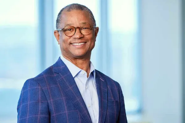 A man wearing a suit and glasses stands in front of large windows