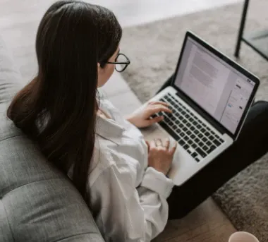 Student with long dark hair and glasses seated on a couch using a laptop