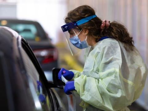 A nurse wearing protective equipment stands beside a car in a parking garage preparing a COVID test swab 
