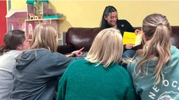 A woman sits on a sofa, reading from a book while showing the pages to a group of girls seated on the floor