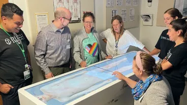 A group of students and teachers gathered around a table with an interactive display screen showing a model of the human body
