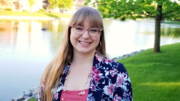 A smiling woman with long hair and glasses stands outdoors in front of a pond