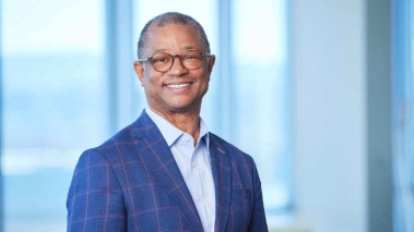Ted Love smiling and wearing a blue suit, standing in front of tall windows