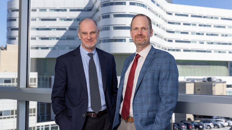 Two men standing together in front of windows with the Rush Tower building visible in the background