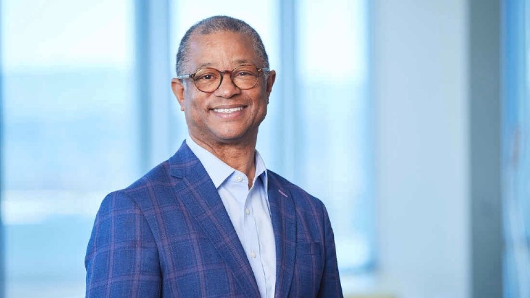 Ted Love smiling and wearing a blue suit, standing in front of tall windows