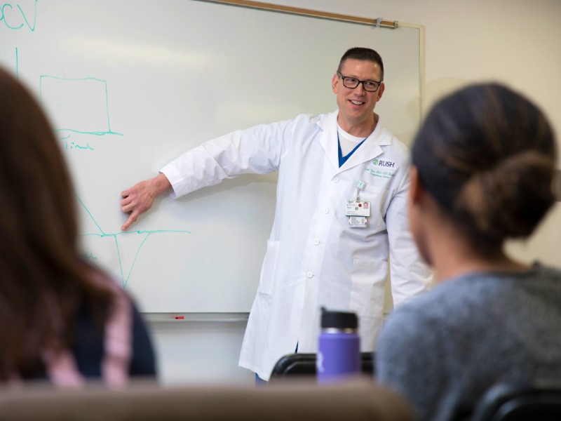 A lecturer in a white coat gestures at a classroom whiteboard